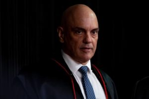 Alexandre de Moraes, no dia de sua posse na presidência do TSE. Ele olha para frente, usando toga, sob a penumbra - Metrópoles