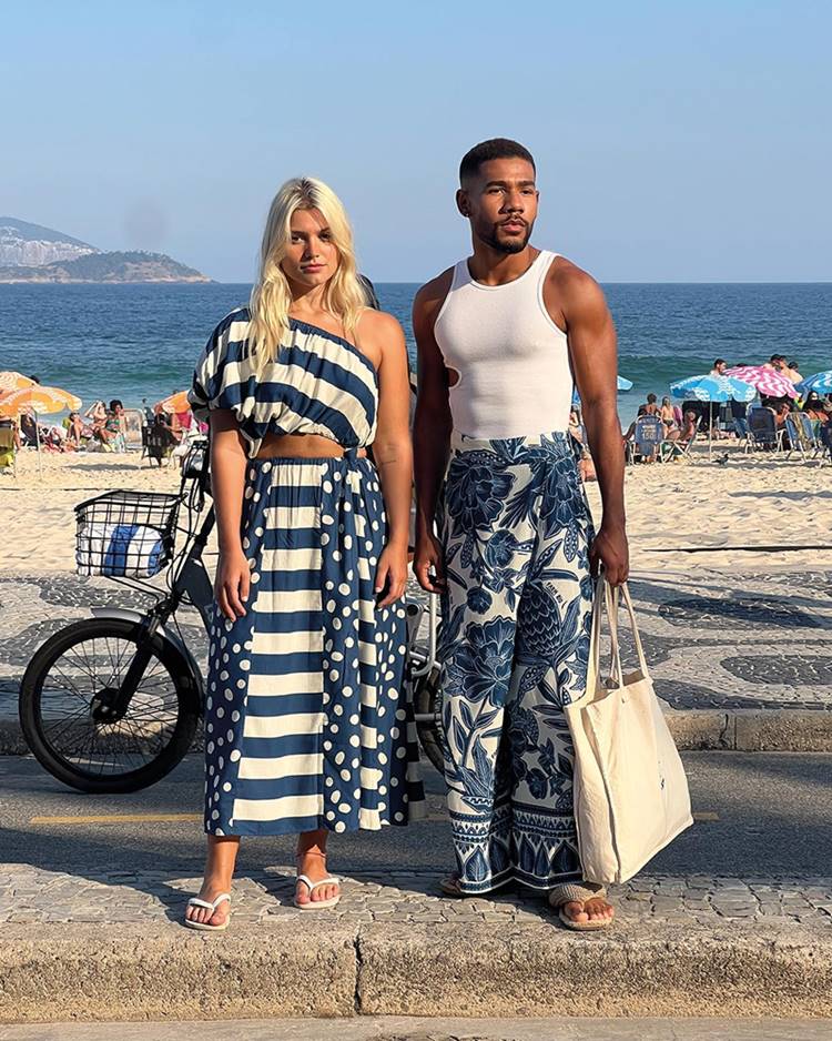 Uma mulher branca, jovem e loira, ao lado de um homem jovem, negro e com o cabelo curto, ambos no calçadão da praia do Rio de Janeiro. Eles usam roupas nas cores azul e branco da marca Farm - Metrópoles 