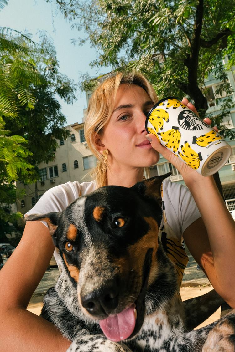 Mulher branca e jovem, de cabelos lisos loiros, posando para foto em um jardim com um cachorro no colo. Ela usa uma camiseta branca e segura um copo térmico branco com desenhos de bananas. A peça é uma parceira entre a marca Farm e a cafetaria Starbucks