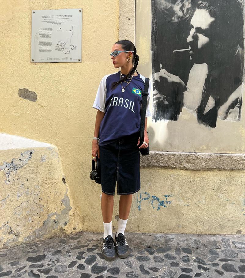 Uma mulher branca e jovem, com cabelo castanho liso amarrado em uma trança, nas ruas de Lisboa, em Portugal. Ela usa óculos escuros, uma camiseta da seleção brasileira de futebol na cor azul, um bermuda preta, meias brancas e um tênis preto da marca Adidas.