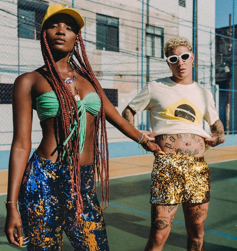 Uma mulher jovem e negra, de cabelos trançados ruivos e longos, posando para foto ao lado de um homem jovem, branco, e de cabelo cacheado loiro. Eles estão em uma quadra de futsal e usam roupas nas cores verde, amarelo e azul, em referência às cores da bandeira do Brasil.