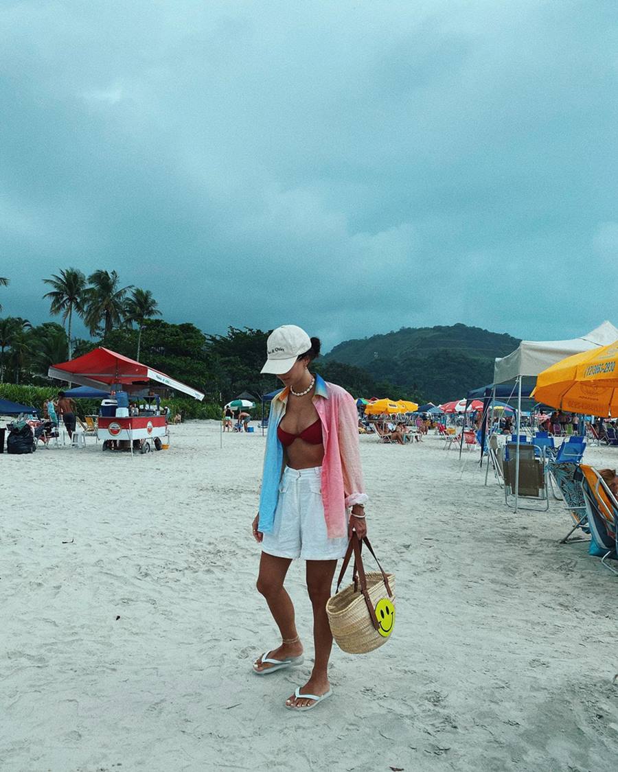 Mulher branca e jovem, de cabelo liso castanho, posando para foto na praia. Ela usa um biquíni vermelho, um shorts branco de linho, uma camisa de botão tingida com cores pastel, boné branco e sandálias Havaianas - Metrópoles