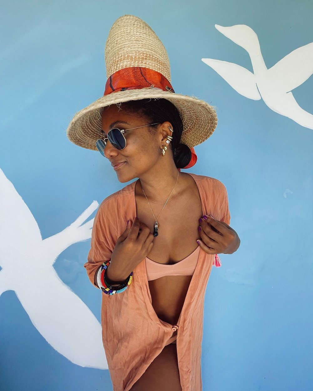 Mulher negra e jovem, de cabelo cacheado amarrado, posando para foto em uma parede azul. Ela usa um biquíni bege, uma camisa de botão bege como saída de praia, óculos escuros e um chapéu de palha - Metrópoles