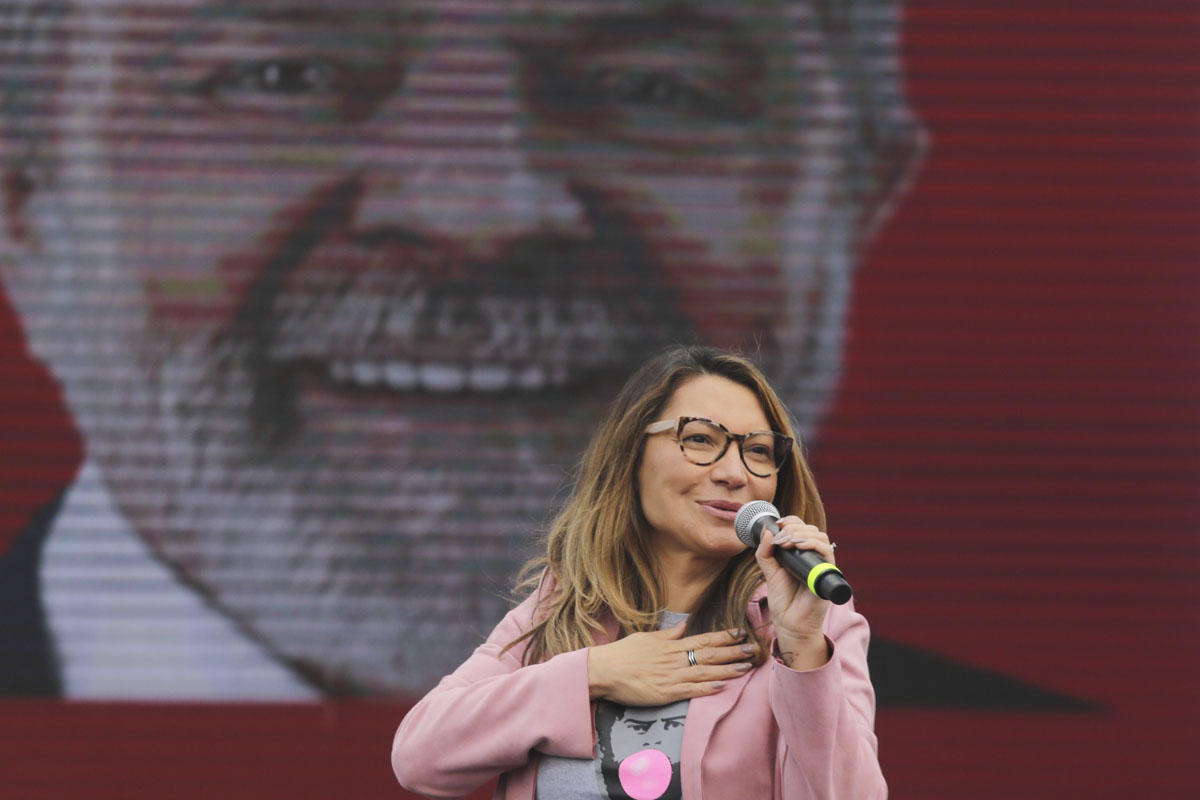 janja de roupa rosa falando ao microfone com imagem de lula ao fundo