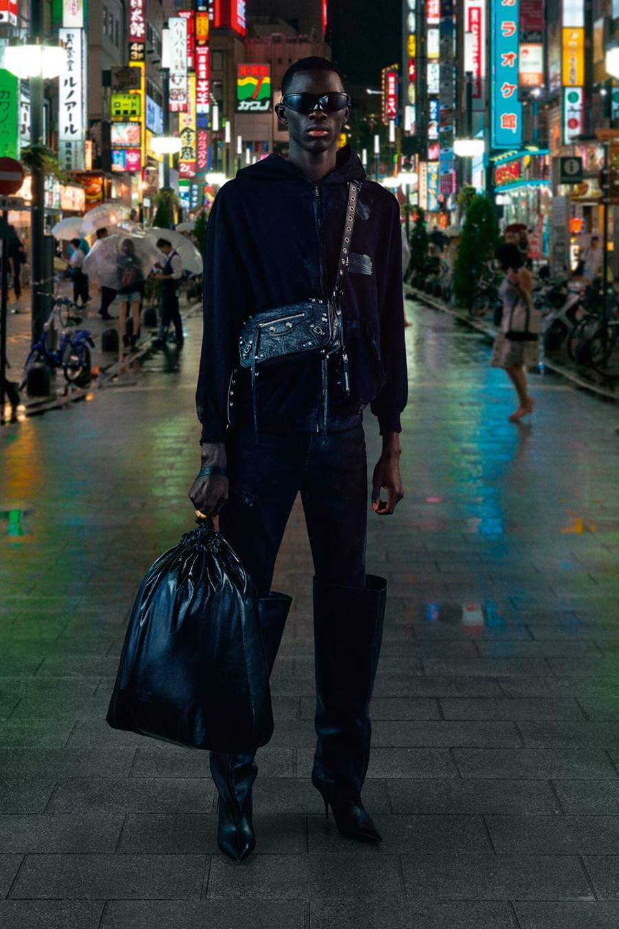 Foto da campanha de outono 2022 da grife de luxo Balenciaga. Na imagem é possível ver um homem negro e jovem em uma rua movimentada. Ele usa um casaco moletom, calça, botas de couro, bolsa curta de couro e óculos, tudo na cor preta. Segura ainda uma sacola preta de lixo. - Metrópoles