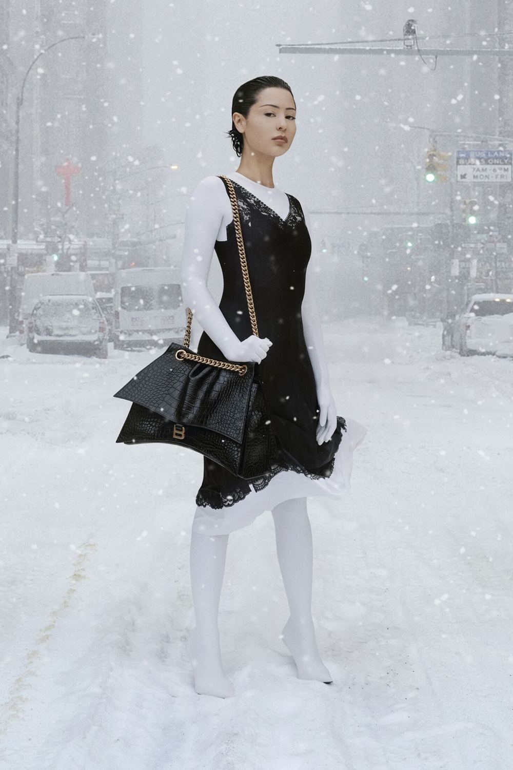 Foto da campanha de outono 2022 da grife de luxo Balenciaga. Na imagem é possível ver uma mulher jovem e branca, de cabelos curtos castanhos, no meio de uma rua coberta por neve. Ela usa um casaco e uma bota, ambos na cor branca, um vestido preto de renda por cima e uma bolsa preta de couro com corrente de metal da marca. - Metrópoles