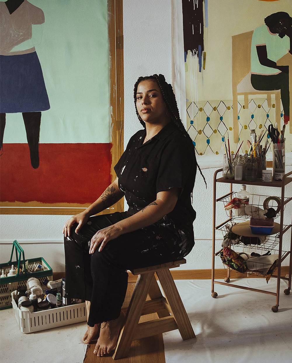 Larissa de Souza, mulher jovem, negra e com cabelos longos trançados, posando para foto no seu ateliê. Usa um macacão preto e, ao fundo, é possível ver quadros pintados por ela e materiais como pincéis e tintas. - Metrópoles