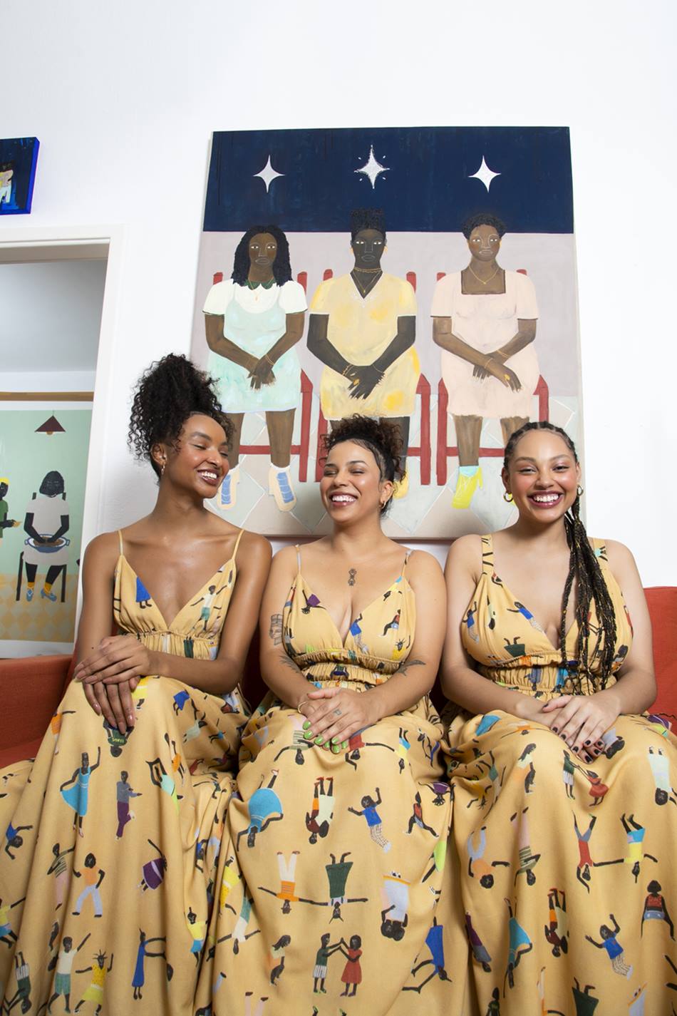 Três mulheres jovem e negras posando para foto com um vestido amarelo longo de alcinha. A peça é fruto de uma coleção especial entre a C&A e a artista plástica Larissa de Souza, a mulher que está sentada no centro. No fundo é possível ver uma obra da artista. - Metrópoles