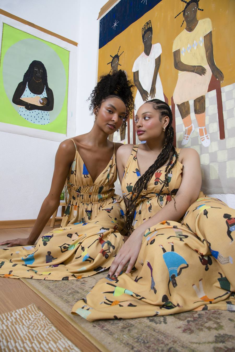 Duas mulheres jovem e negras, uma com cabelo cacheado e outra com tranças, posando para foto com um vestido amarelo de alça. A peça é fruto de uma coleção especial entre a C&A e a artista plástica Larissa de Souza. No fundo é possível ver uma obra da artista. - Metrópoles