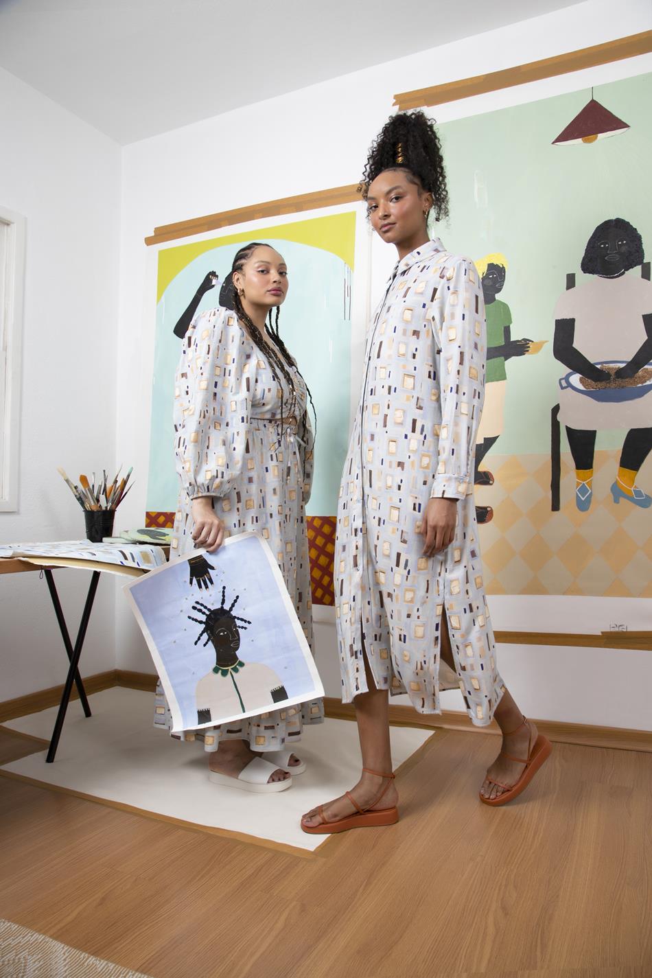 Duas mulheres jovem e negras, uma com cabelo cacheado e outra com tranças, posando para foto com um vestido e um conjunto de saia e blusa. As roupas são azuis claras, possuem pequenos desenhos colorudos e são fruto de uma coleção especial entre a C&A e a artista plástica Larissa de Souza. No fundo é possível ver uma obra da artista. - Metrópoles