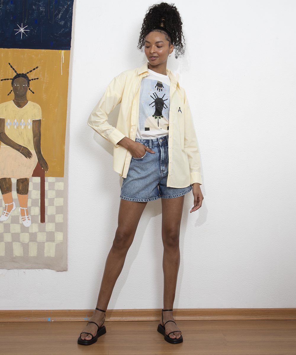 Modelo jovem e engra, com cabelo cacheado amarrado em um rabo de cavalo, posando para foto em um fundo branco com uma obra de arte ao fundo, pendurada na parede. Ela usa um shorts jeans, uma camiseta branca e uma camisa amarela de botão por cima, aberta, peças fruto da coleção especial entre a C&A e a artista plástica Larissa de Souza. - Metrópoles