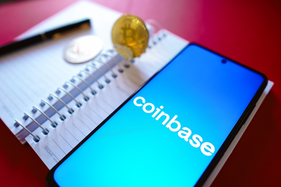 Foto colorida. Caderno com uma caneta, lápis e uma moeda. Um celular está ligado com o nome Coinbase - Metrópoles