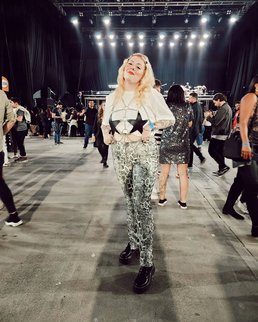 Mulher branca e jovem, de cabelo liso ondulado, posando para foto em frente ao palco de um show em um estádio. Ela usa uma camiseta branca, uma calça de couro com estampa de cobra, um coturno preto e batom vermelho. - Metrópoles