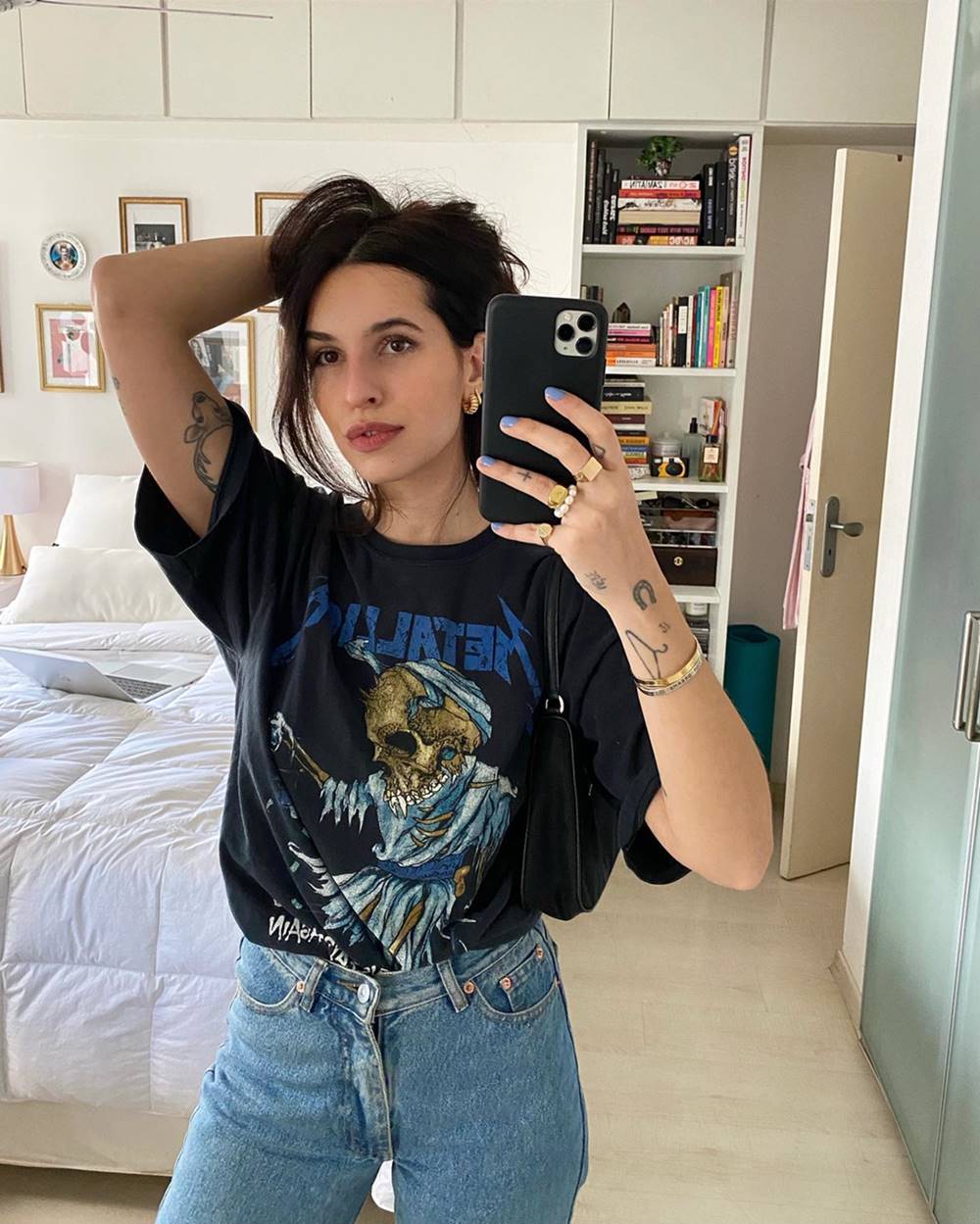 Mulher branca e jovem, de cabelo castanho liso e longo, posando para foto em frente ao espelho no próprio quarto. É possível ver, no fundo, uma cama e uma estante com livros. Ela usa uma camiseta cinza com estampa do Metallica e calça jeans