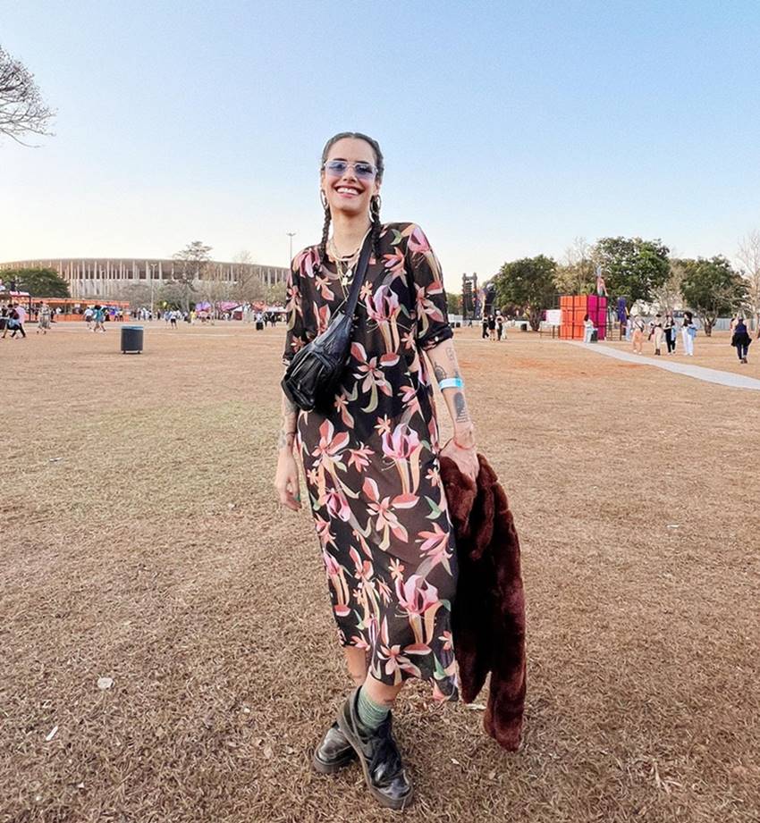 Mulher morena e jovem, de cabelos trançados, posando para foto no festival COMA, em Brasília. Ela usa um vestido preto com flores rosas, um casaco de pele marrom, um óculos escuros e uma pochete preta de couro. - Metrópoles