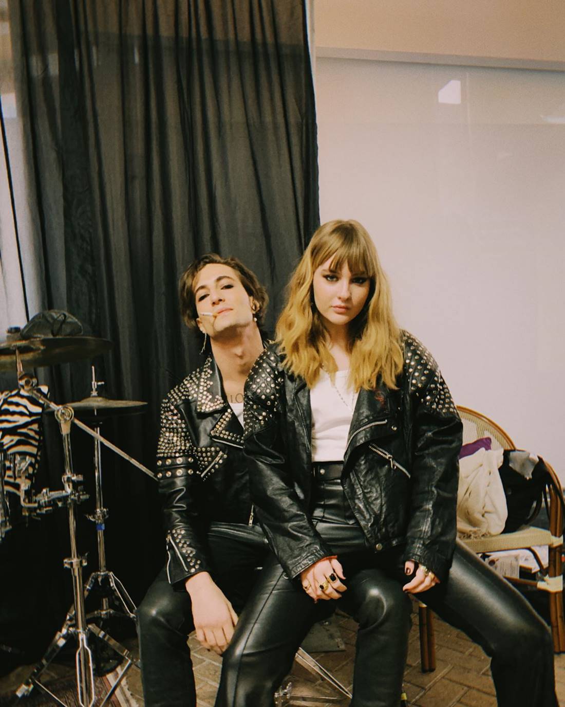 Mulher branca e jovem, de cabelo liso loiro, posando para foto sentada no colo de um homem branco, jovem, de cabelo preto liso grande. Eles são da banda de rock Måneskin. Os dois usam looks iguais: camisa branca e calça + jaqueta de couro preto. - Metrópoles