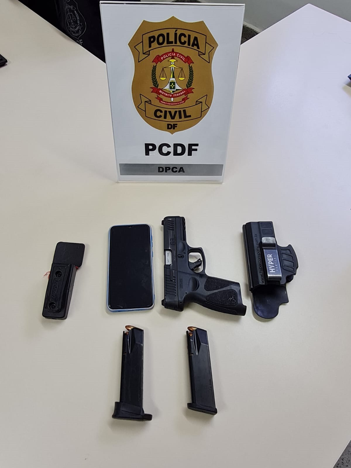 Conjunto de duas armas expostas em mesa branca com logo da Polícia Civil do Distrito Federal atrás. Foto colorida