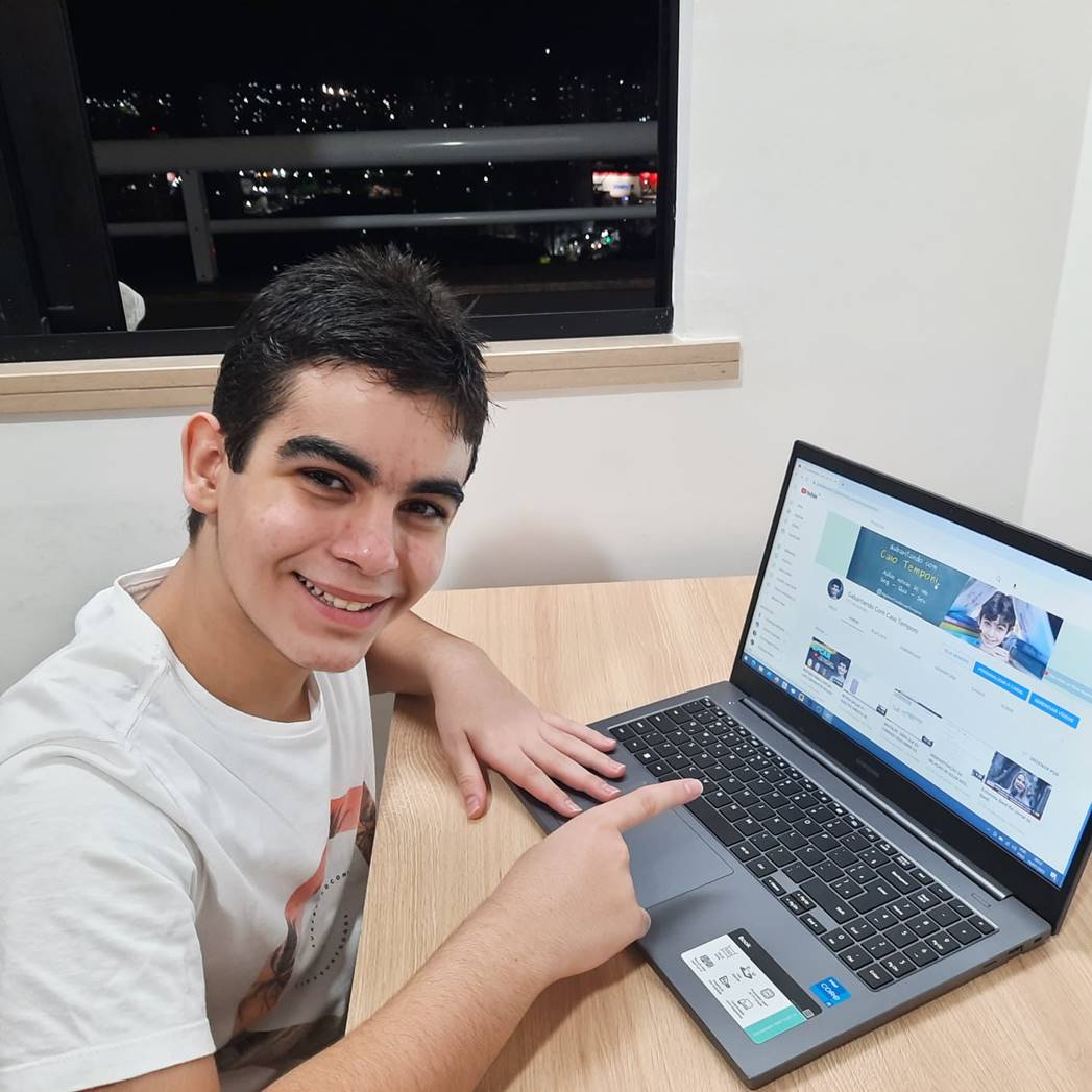 Menino de camisa branca e sentado na cadeira enquanto usa notebook