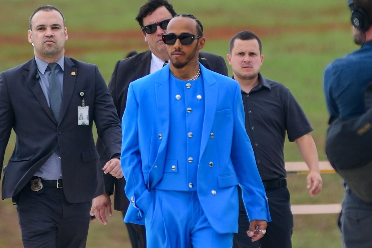 Chegada do piloto britânico Lewis Hamilton na Câmara dos Deputados para receber título de cidadão honorário do Brasil. Ele está cercado de uma comitiva e usa terno azul celeste. Ele faz um sinal de joia com a mão - Metrópoles