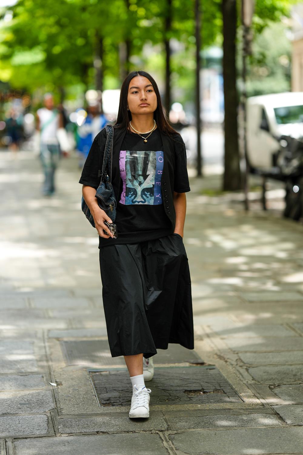 Mulher negra e jovem, de cabelos lisos, durante a Paris Fashion Week - Menswear Spring/Summer 2023, em 26 de junho de 2022 em Paris, França. Ela usa um colar de pérolas brancas, um colar de corrente de ouro, brincos de ouro, uma camiseta preta com estampa estampada, uma bolsa de ombro jeans azul marinho, calça preta de pernas largas, meias brancas, tênis de couro branco da Adidas. - Metrópoles