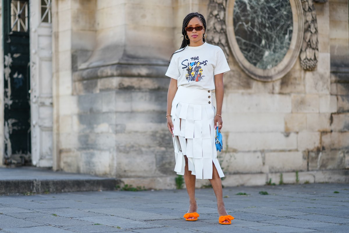 Mulher negra e jovem, de cabelo longo trançado, durante uma sessão de fotos de moda de rua, em 11 de maio de 2022 em Paris, França. Ela usa óculos escuros marrons, brincos dourados, camiseta branca com preto e azul e amarelo com estampa dos Simpsons, pulseiras douradas, saia midi cintura alta branca com franjas, bolsa Saddle pequena azul neon brilhante da Dior, uma Juste dourada Pulseira Un Clou da Cartier. - Metrópoles