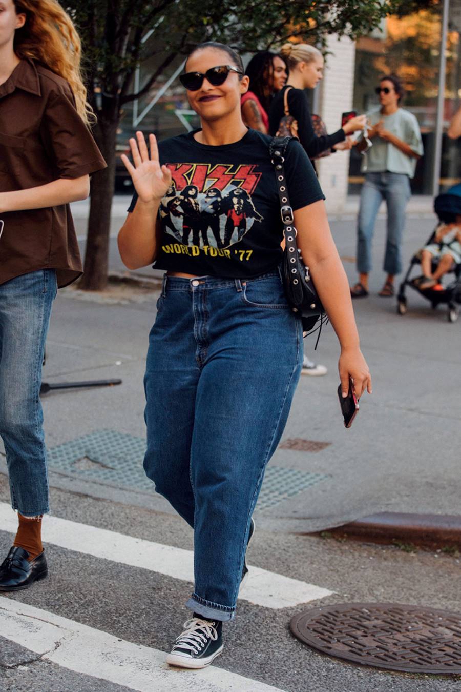 Mulher negra e jovem, de cabelo preto liso amarrado, andando pelas ruas de Nova York, nos EUA. Ela usa uma camiseta preta com estampa da banda Kiss, uma calça jeans, tênis All Star Converse preto, óculos escuros e uma bolsa preta de couro da marca Balenciaga. - Metrópoles