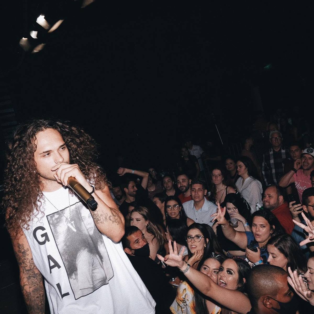 O cantor Vitão, um homem jovem, moreno, com cabelo cacheado castanho e longo, segurando o microfone e se apresentando em um teatro fechado. Usa uma camiseta branca com foto em preto e branco da cantora baiana Gal Costa.