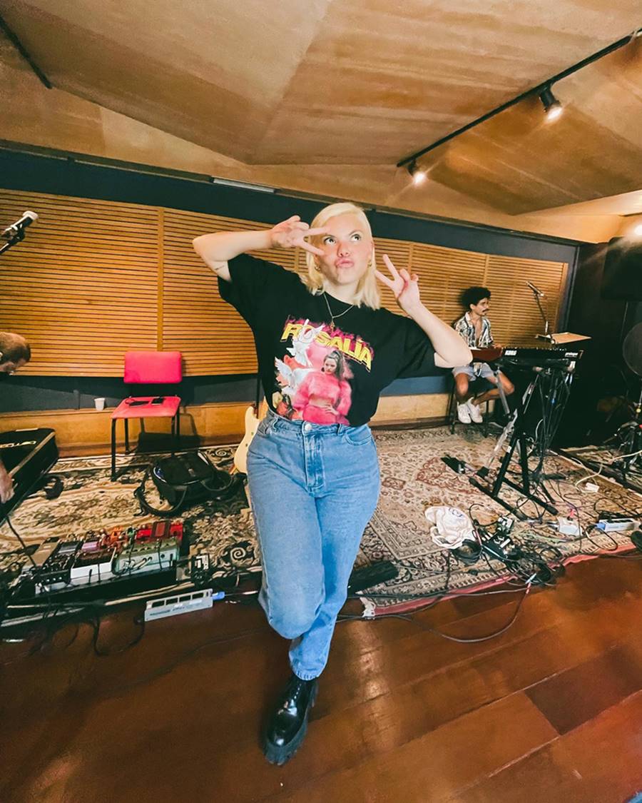 A cantora Duda Beat, uma mulher branca, jovem e com cabelo liso loiro, posando para foto em um estúdio de música. Usa calça jeans, bota preta de couro e uma camiseta preta com estampa da cantora Rosalía. - Metrópoles