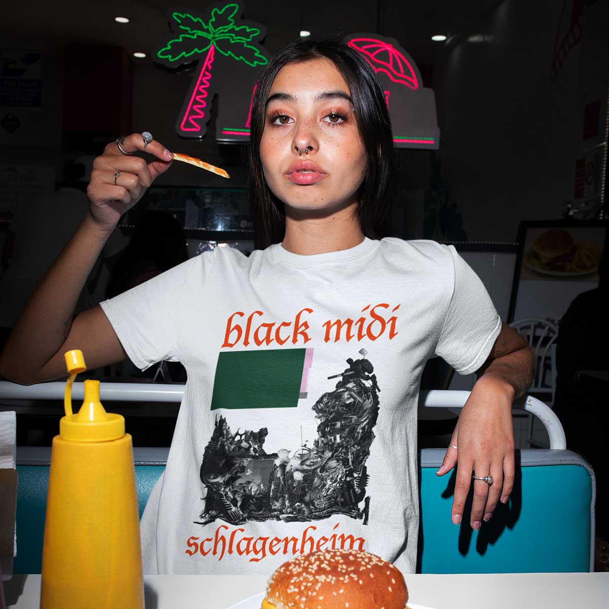 Mulher jovem e morena, de cabelo liso, posando para foto na mesa de uma lanchonete de fast food. Segura batatas fritas e um hamburger. Usa uma camiseta branca com estampada da banda de rock britânica Black Midi, que foi fundada em 2017.