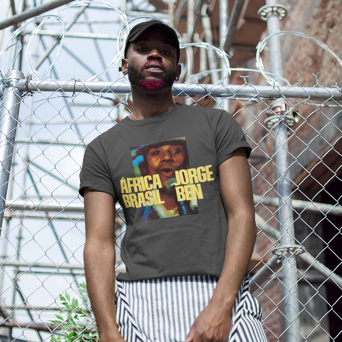 Homem jovem, negro e de barba pintada de vermelha, posando para foto em frente a uma grade prateada. Ele usa um boné preto e uma camiseta cinza com estampa do cantor Jorge Ben Jor.