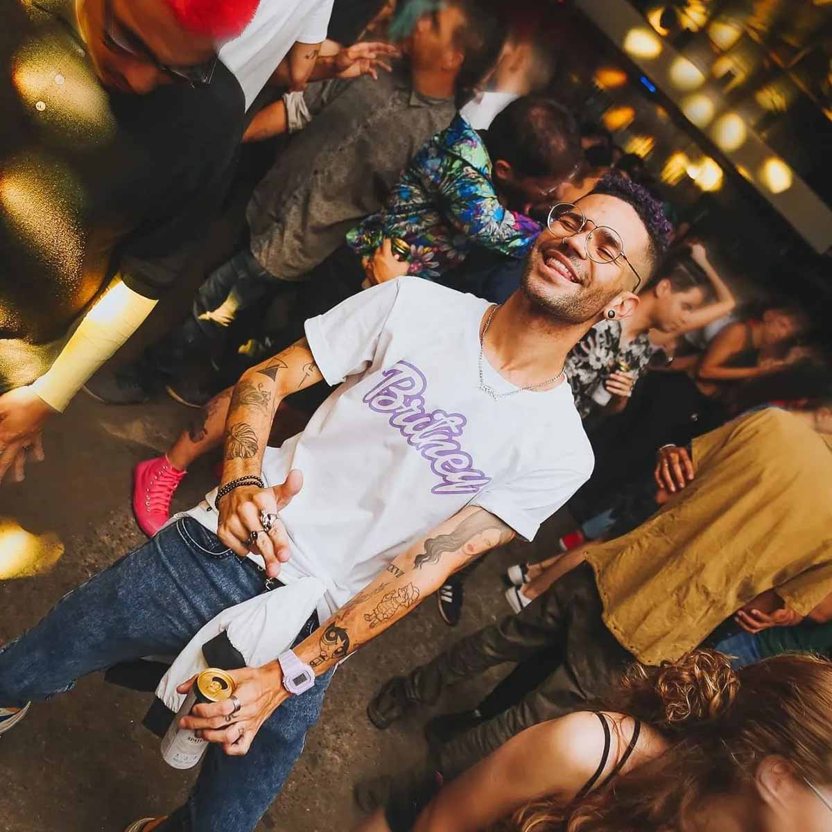 Homem negro e jovem, de cabelo castanho curto, posando para foto em uma festa. Ele usa uma camiseta branca com o nome da cantora Britney Spears estampado.