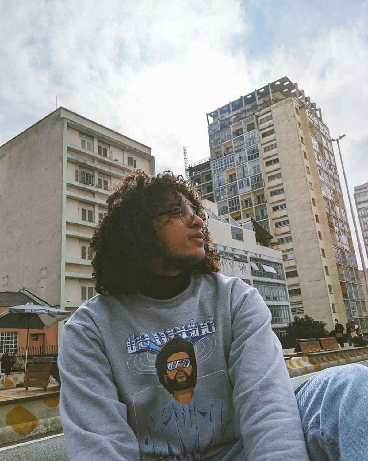 Homem jovem e negro, de cabelo longo cacheado, posando para foto em meio a prédios de São Paulo. Usa um moletom cinza com a estampa do cantor The Weekend.