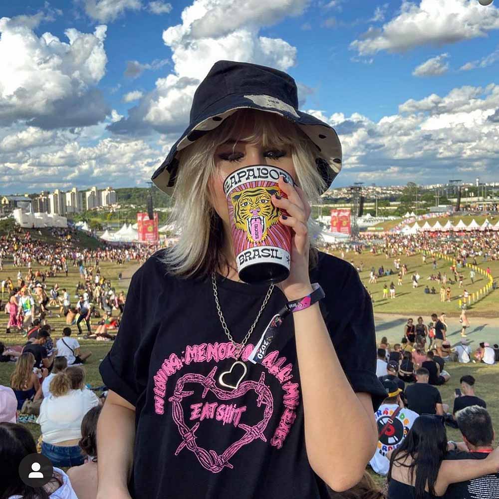 Mulher jovem, branca e loira posando para foto no festival Lollapalooza. Ela usa uma camiseta preta e um chapéu estilo pescador também preto. Está segurando um copo estampado e bebendo.