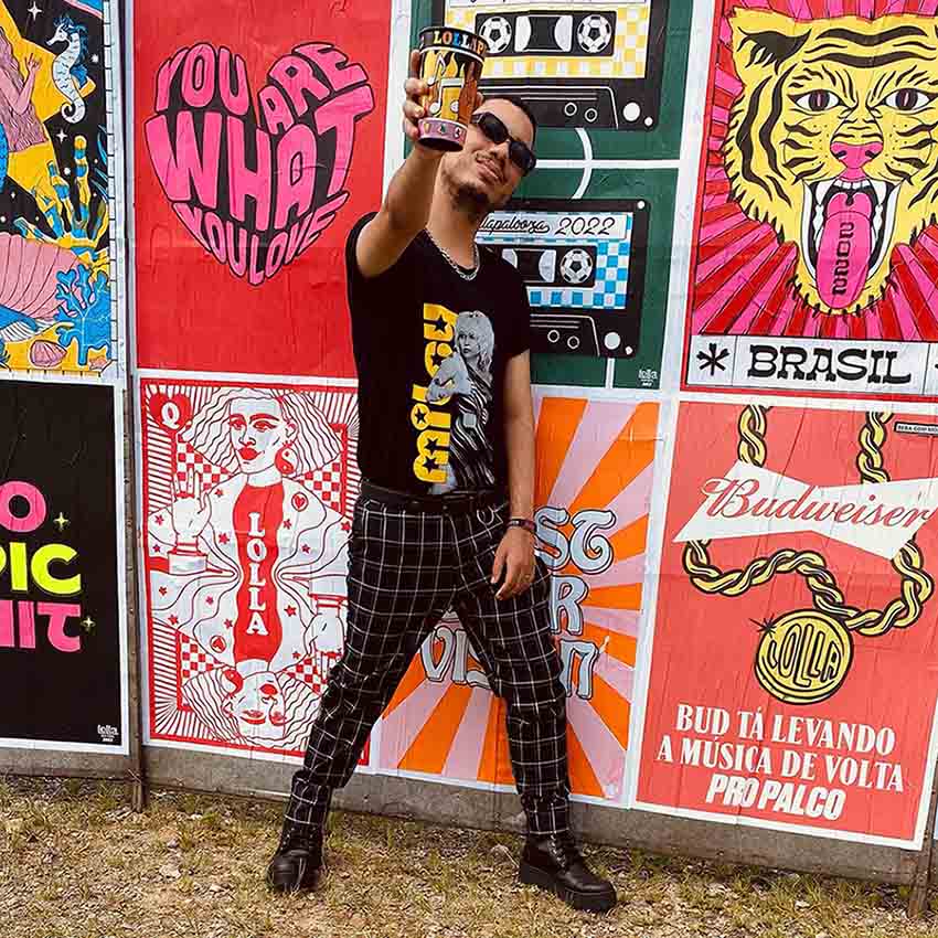 Homem jovem, negro e de cabelo curto cacheado posando para foto no festival Lollapalooza. Ele usa uma camiseta preta com estampa da cantora Miley Cyrus, calça listrada preta e branca, bota de couro preta e óculos escuros.