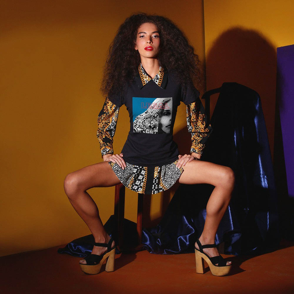 Mulher branca e jovem, de cabelo cacheado longo e volumoso, posando para foto em um fundo amarelo e sentada em um banquinho de madeira. Usa uma saia preta e branca, uma camiseta preta com estampa da Gal Costa e uma sandália de salto de madeira.