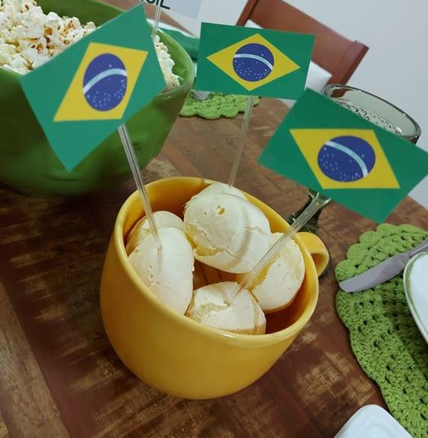 Foto colorida de uma xícara amarela com pão de queijo dentro e pequenas bandeirinhas do Brasil 