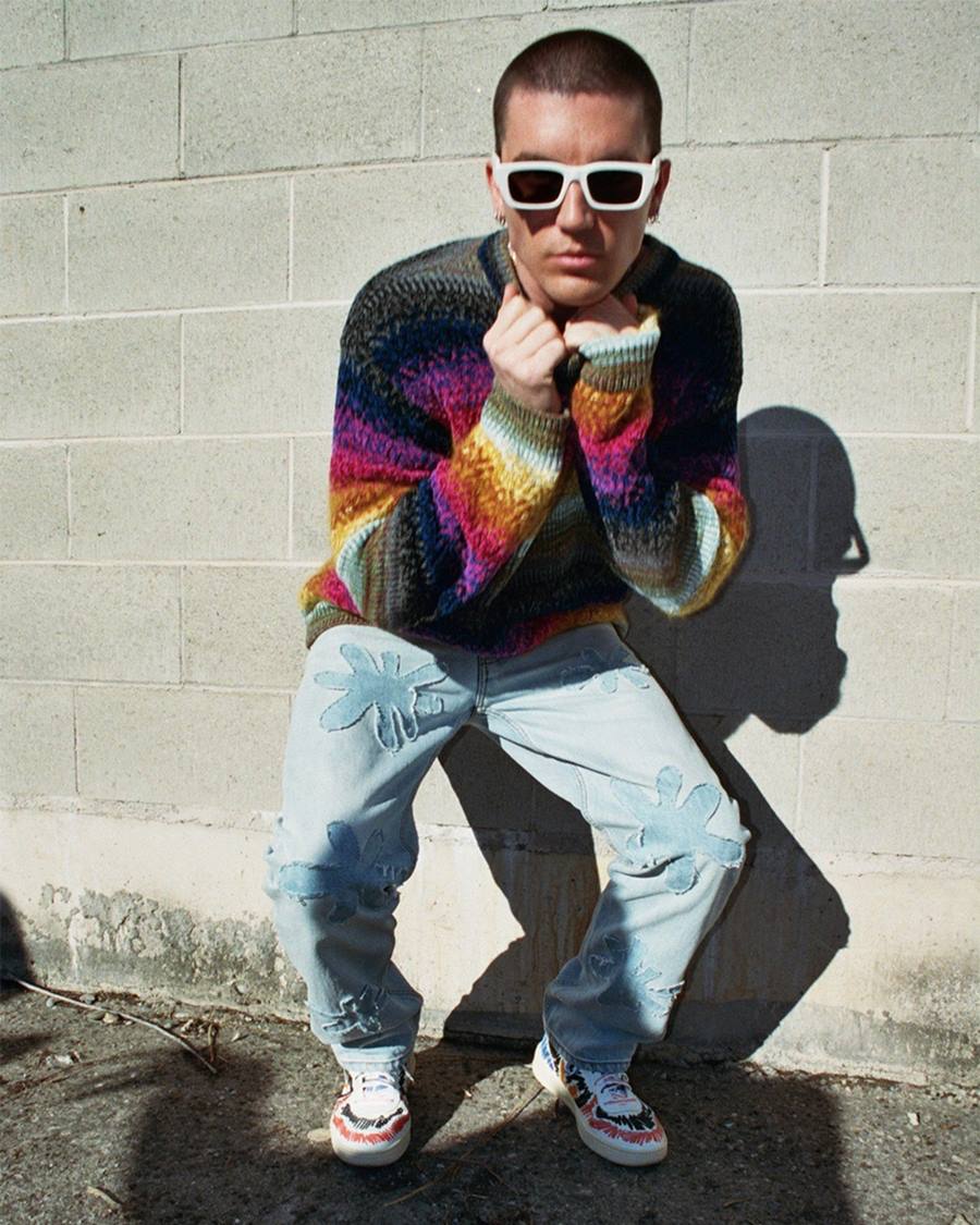 Homem branco e jovem, com cabelo curto preto, posando para foto em frente a um muro de tijolos brancos. Usa uma calça jeans, um suéter listrado colorido e um óculos escuros de armação branca.