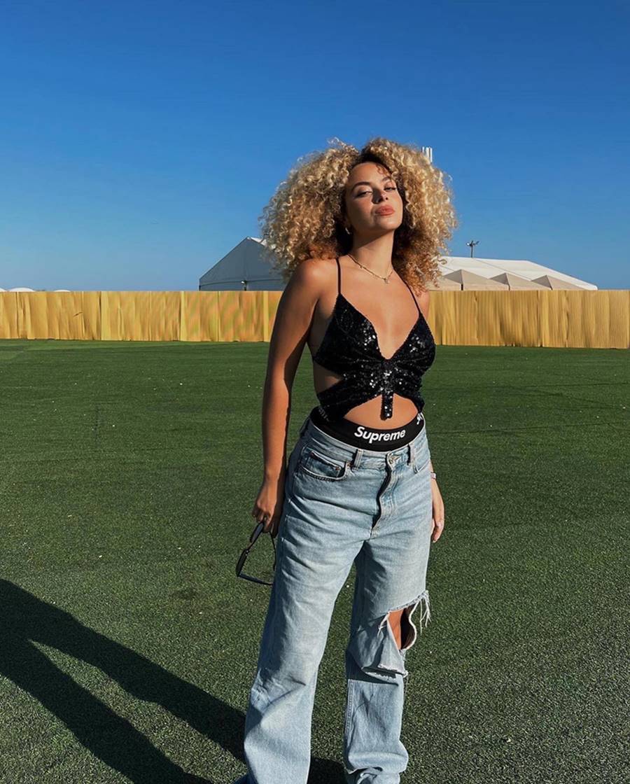 A influenciadora digital Julia Rodrigues, uma mulher jovem, negra, com cabelo cacheado longo e loiro, posando para foto no festival de música Rock in Rio. Ela veste um top preto com recortes de lantejoulas e uma calça jeans folgada com rasgo no joelho. - Metrópoles