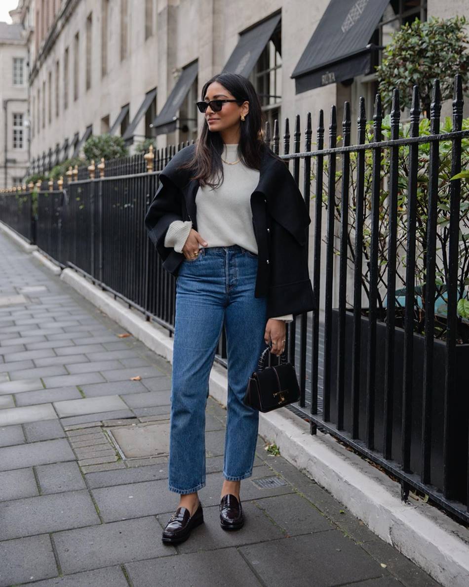 A influenciadora digital Hannah, uma mulher jovem, morena, com cabelo liso longo, posando para foto em frente a uma grade preta de ferro. Ela veste um casaco cinza e outro preto por cima, calça jeans, sapato mocassim preto de couro e óculos de sol. - Metrópoles