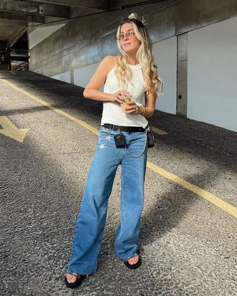 A influenciadora digital Larissa Proença, uma mulher jovem, branca, com cabelo loiro longo e ondulado, posando para foto em um estacionamento. Ela veste uma camiseta branca, calça jeans azul, óculos escuros e uma sandália preta. - Metrópoles