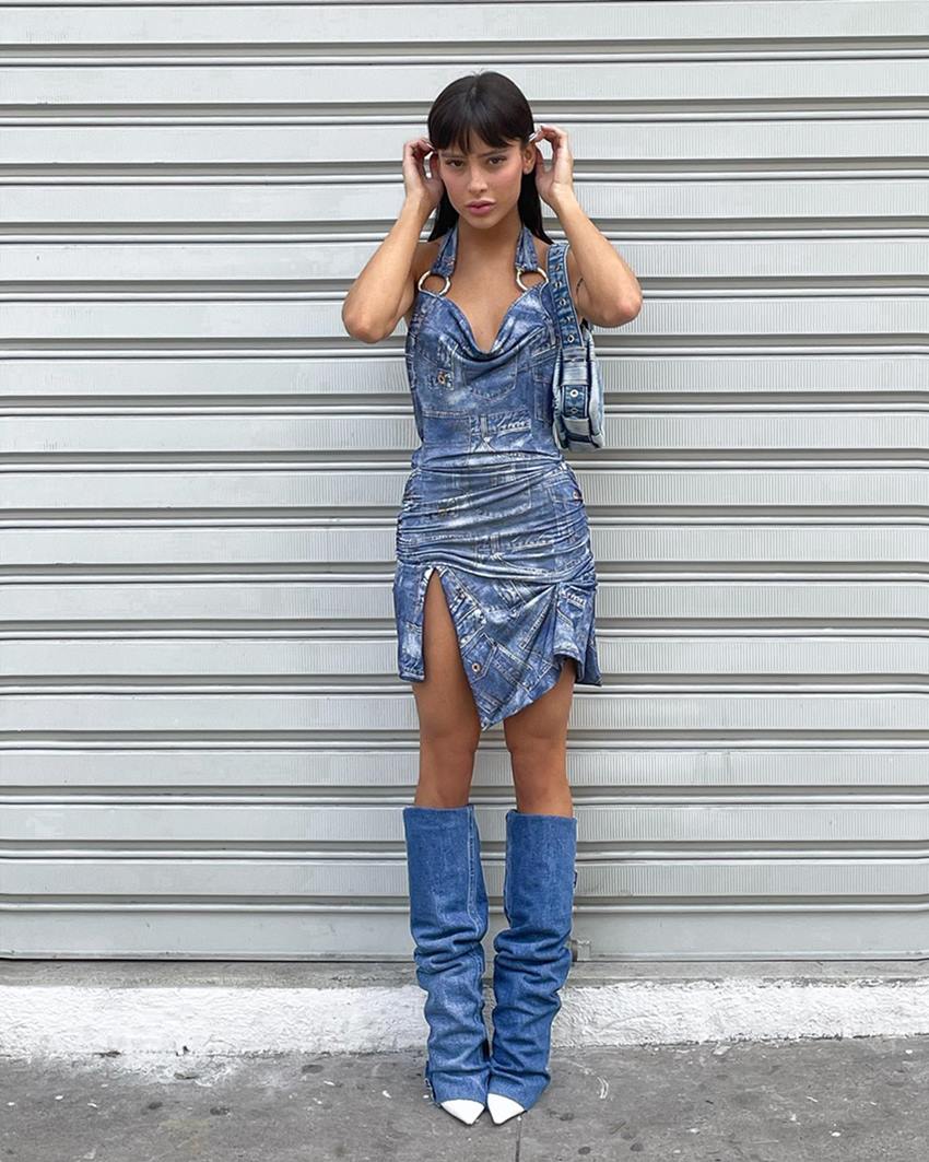 A influenciadora digital Livia Marques, uma mulher jovem, branca, com cabelo castanho longo e liso, posando para foto em um estacionamento. Ela um vestido jeans, uma bolsa jeans e uma bota no mesmo tecido. - Metrópoles