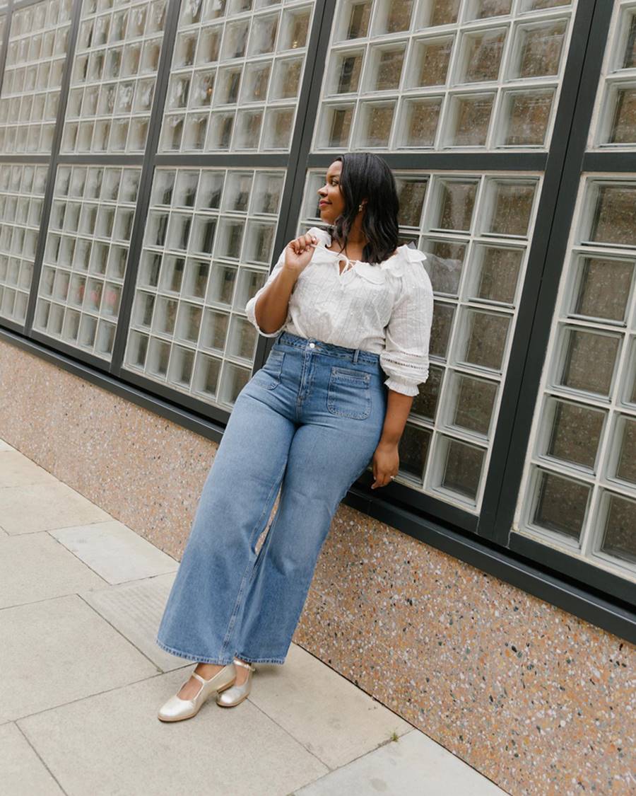 A influenciadora digital Karina, uma mulher jovem, negra, com cabelo castanho ondulado, posando para foto em um estacionamento. Ela um calça jeans folgada, uma blusa branca de babado e uma sapatilha dourada. - Metrópoles