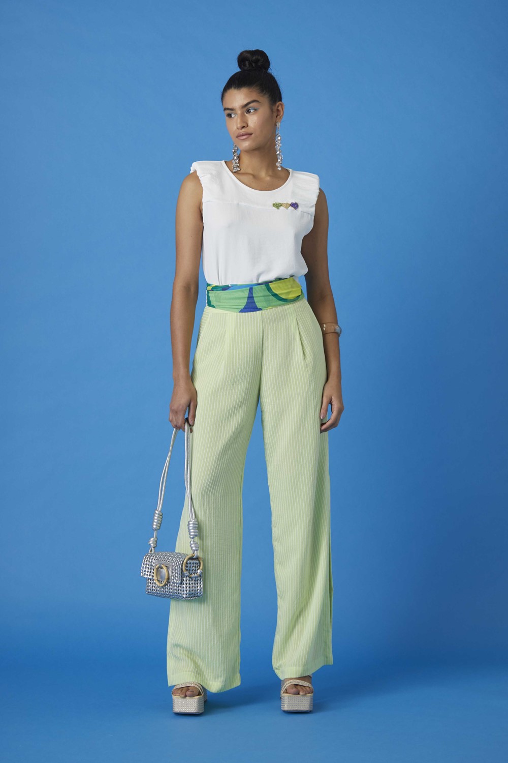 Mulher morena e jovem, de cabelo castanho liso amarrado em um coque, posando para foto em um fundo azul liso. Ela usa uma camiseta branca, uma calça pantalona verde clara, sandália de salto e uma bolsa prata de brilho.