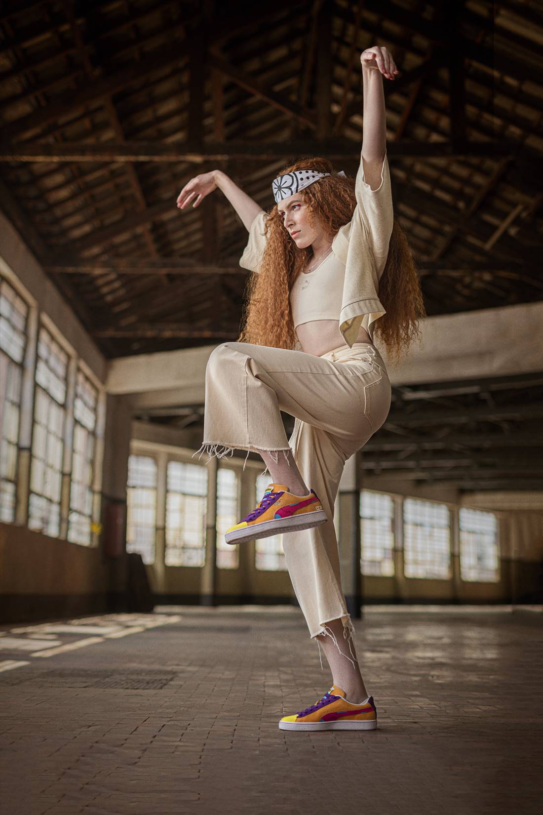 Mulher jovem e branca, de cabelo ruivo cacheado, posando em campanha da marca Puma. Ela usa calça e blusa, ambas na cor bege, e um tênis colorido.