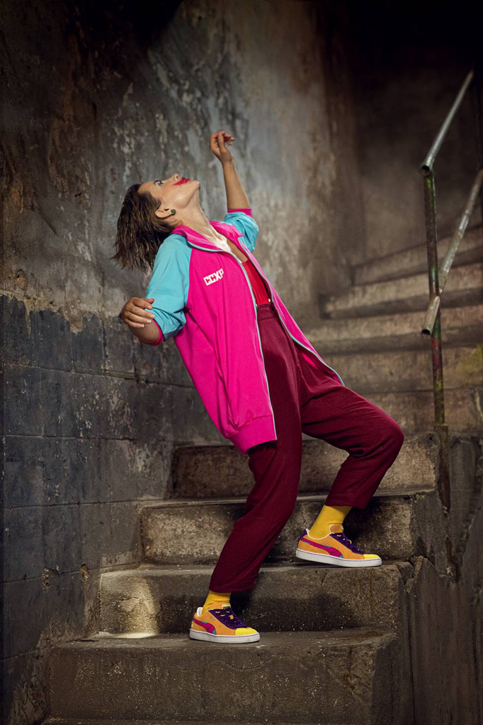 Mulher jovem e branca, com cabelo loiro curto, posando para foto em campanha da marca Puma. Usa uma calça cor de vinho, um casaco rosa e um tênis amarelo. - Metrópoles