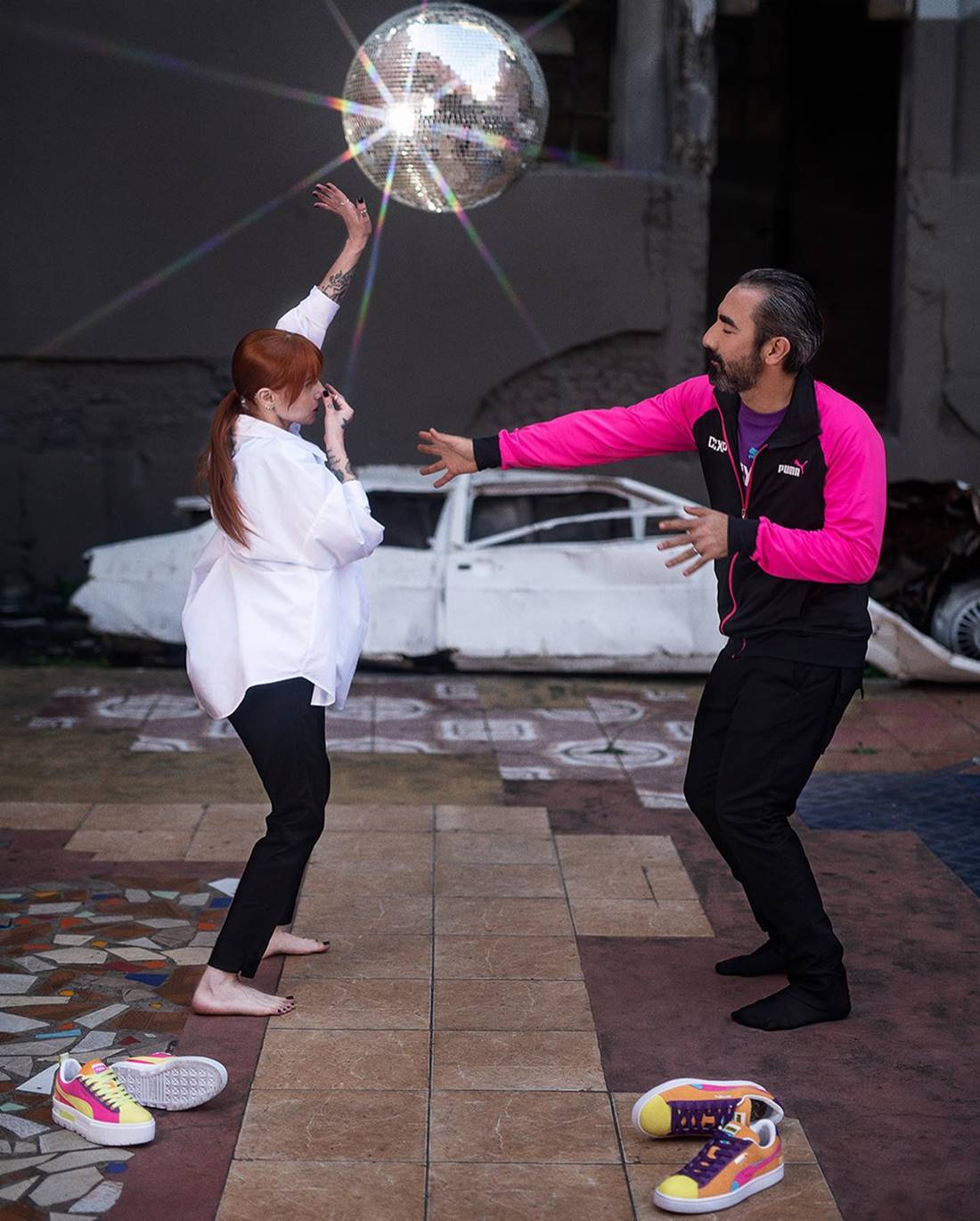 Homem e mulher simulam dançar em uma pista com globo de luz. A foto imita a cena do filme Pulp Fiction e é parte de uma campanha publicitária da marca Puma. A mulher é ruiva e branca, com camisa branca e calça preta, e o homem usa uma roupa toda preta. - Metrópoles