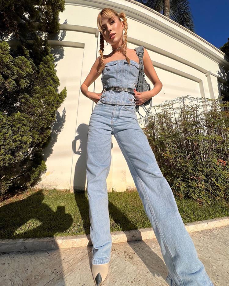 Uma mulher jovem, branca e loira posa para foto em frente a uma casa na capital paulista. Ela usa um top sem alça, uma calça estilo wide leg e uma bolsa da marca Balenciaga. Todas as peças são no tecido jeans. - Metrópoles