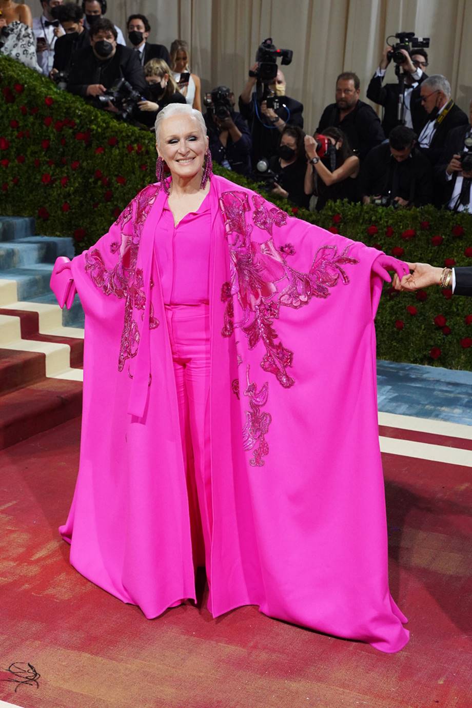 A atriz Glenn Close, uma mulher branca, idosa e com cabelos brancos, no tapete vermelho do baile Met Gala de 2022. Ela usa blusa de botão, calça de alfaiataria, sandália plataforma e uma capa, tudo em um tom de rosa choque. As peças são da marca Valentino. - Metrópoles