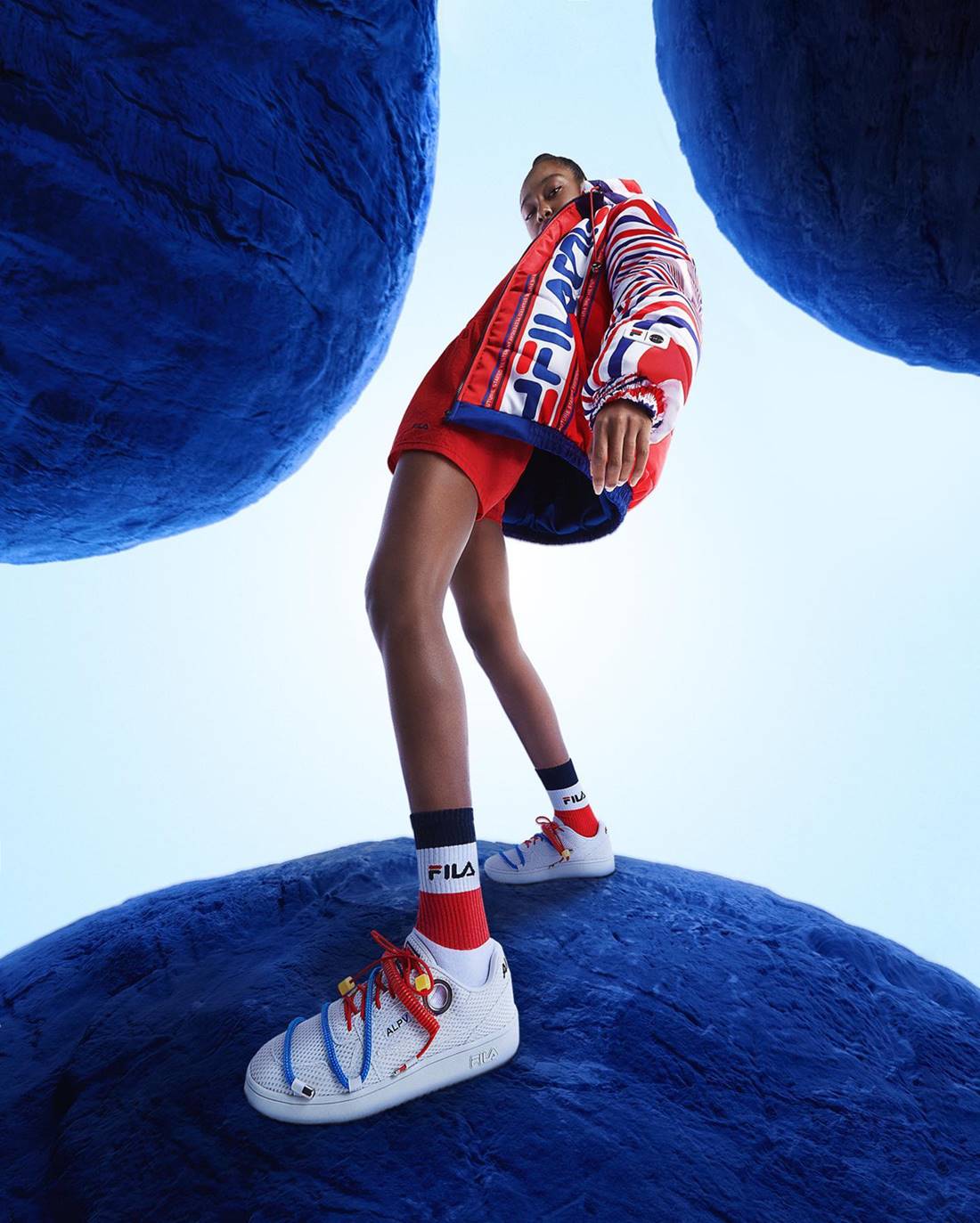 Foto da campanha de divulgação da Fila com o Alexandre Pavão. Na imagem é possível ver uma modelo negra e jovem, de cabelo amarrado, usando shorts e camisa vermelhos e um casaco com a logo da Fila em branco, vermelho e azul. Usa ainda um tênis branco. - Metrópoles.