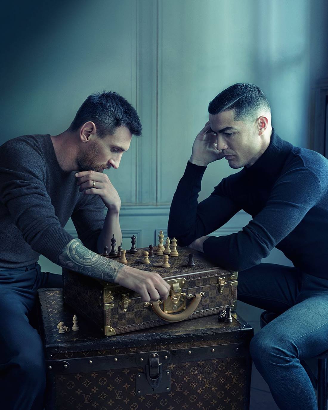 Os jogadores Lionel Messi e Cristiano Ronaldo em campanha de divulgação da grife Louis Vuitton. Estão sentados em bancos e jogam xadrez. O tabuleiro de xadrez está em cima de dois baús da Louis Vuitton. - Metrópoles